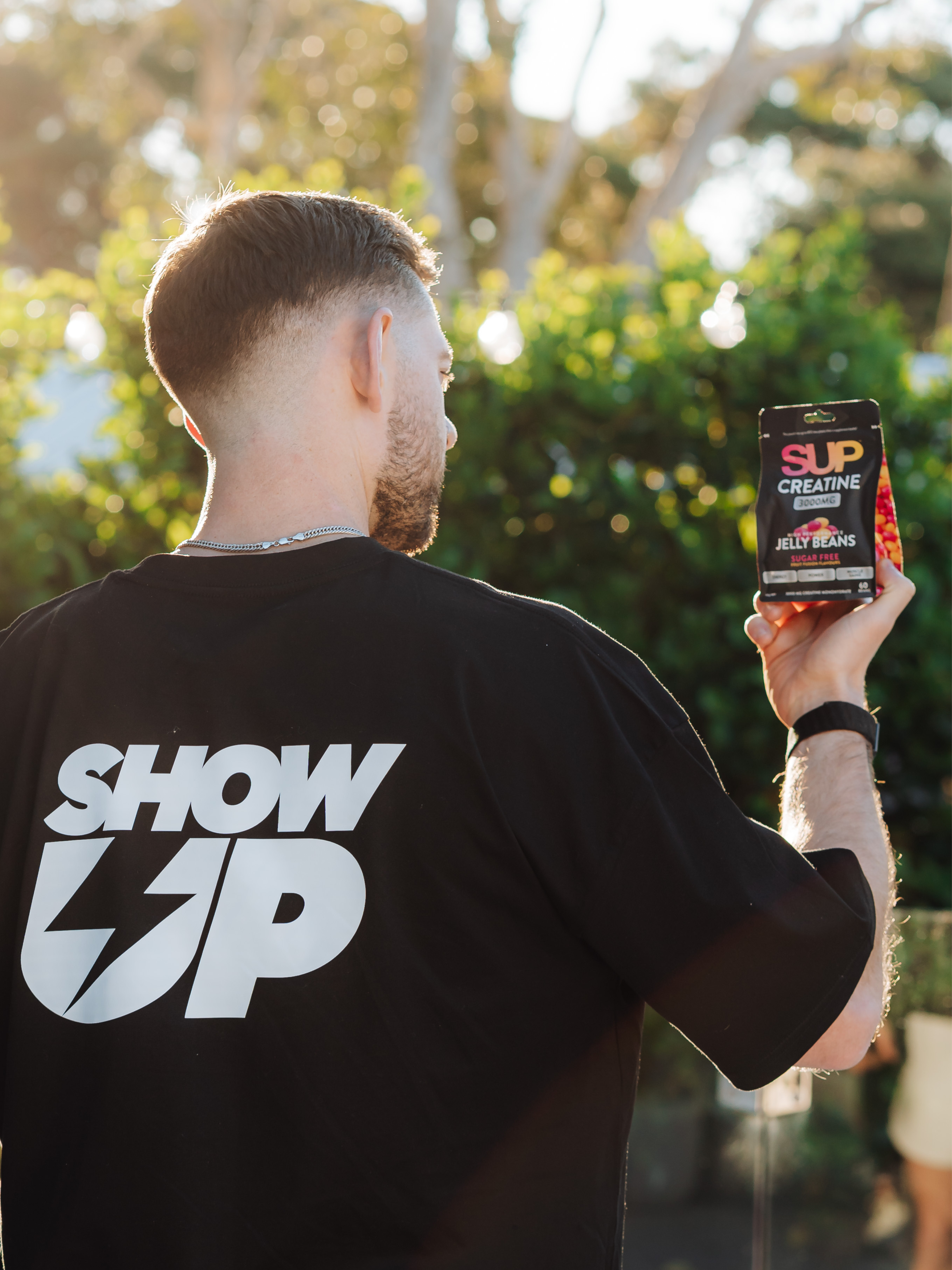 A man seen from behind wearing a black t-shirt with a large white 'SHOW UP' logo on the back, holding up a black supplement bottle labeled 'SUP CREATINE' in an outdoor setting with green foliage in the background.