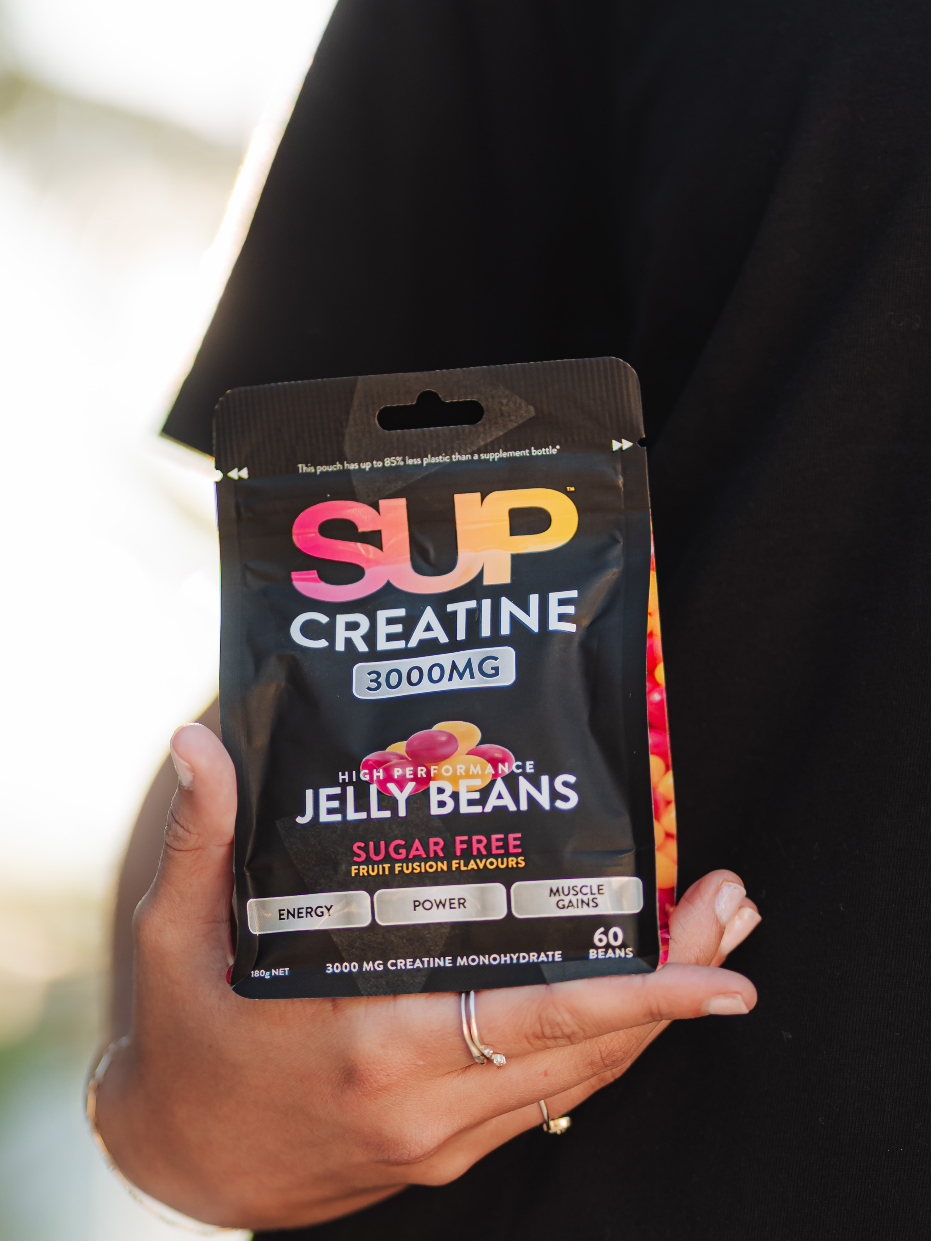 Image of A hand holding a black package of SUP Creatine 3000MG Jelly Beans, with a person wearing a black shirt in the background.